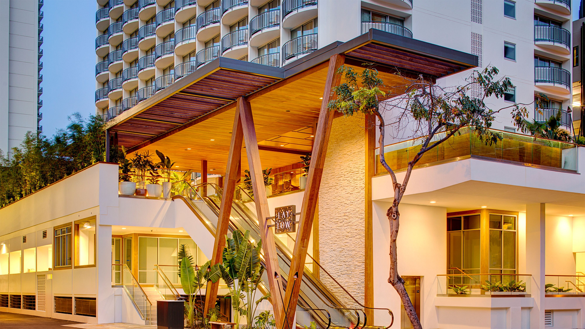The image shows a modern building with a distinct V-shaped wooden support and glass accents, surrounded by greenery and featuring multiple balconies.