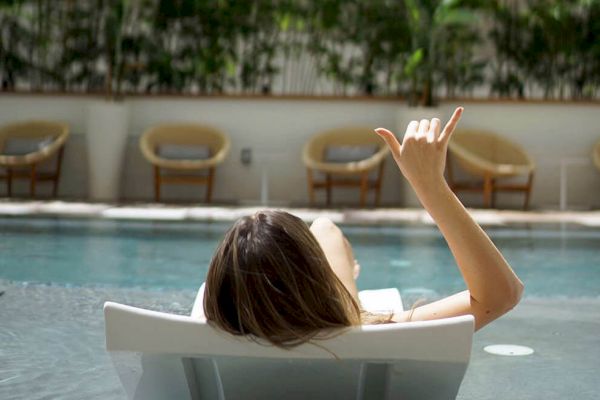 A person is relaxing on a lounge chair by a pool, making a hand gesture, with chairs and plants in the background.