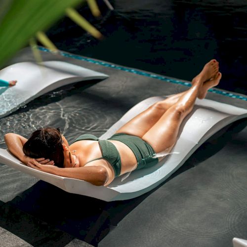 A person in a swimsuit lounges on a chair partially submerged in water, surrounded by a relaxed outdoor setting with greenery.