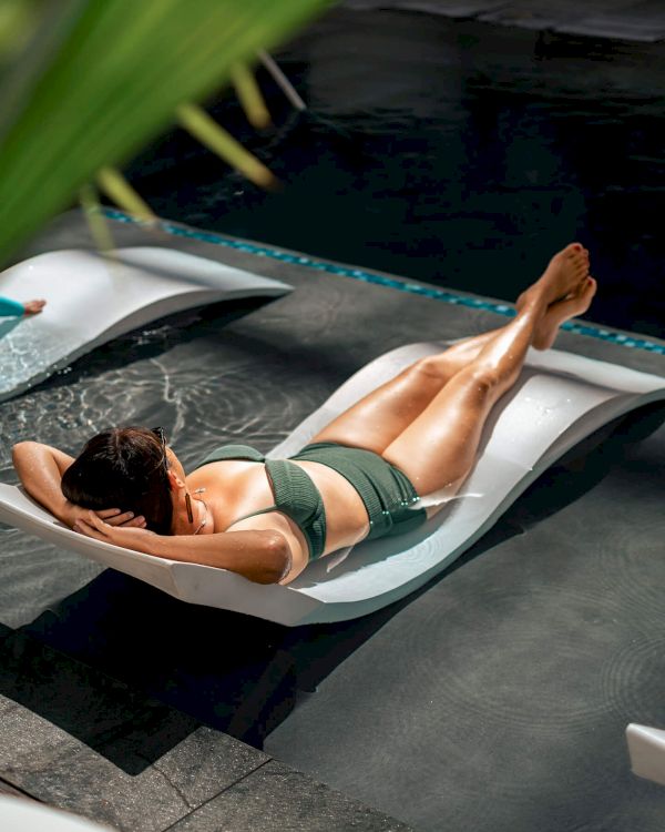 A person in a swimsuit lounges on a chair partially submerged in water, surrounded by a relaxed outdoor setting with greenery.