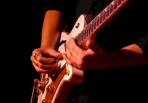 A person playing an electric guitar, focusing on their hands and the instrument in a dimly lit setting.