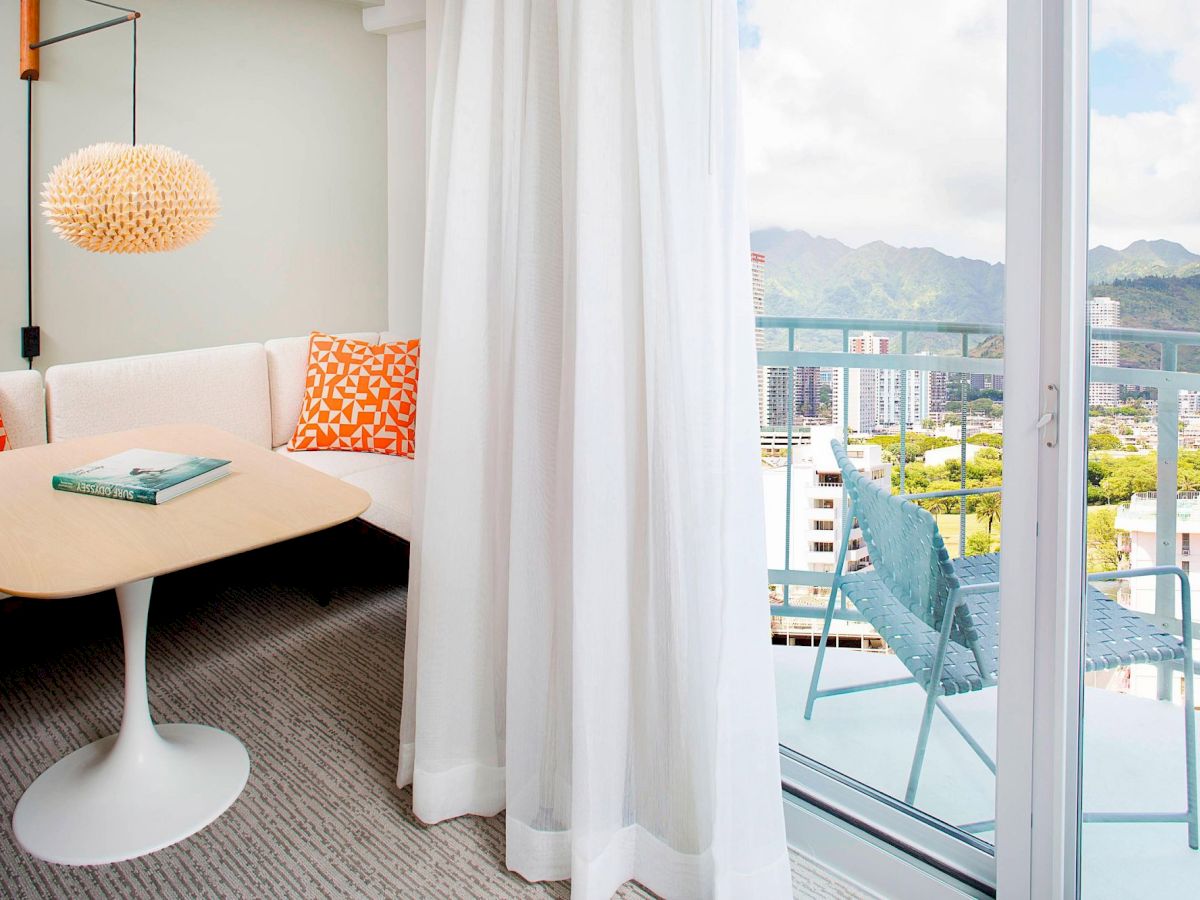 A cozy room with a table, couch, and pendant lamp overlooks a balcony with seating, offering a scenic city and mountain view through glass doors.
