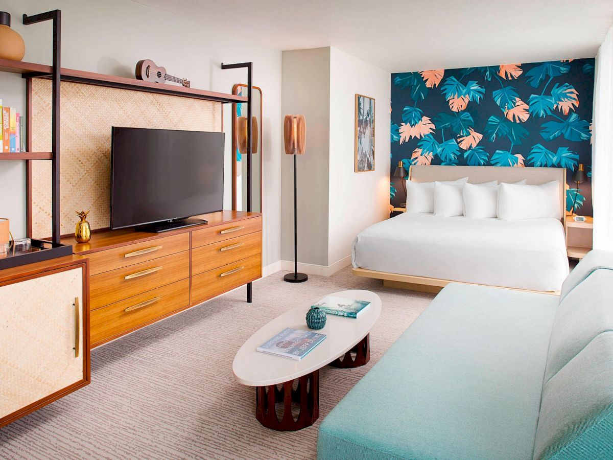 A modern hotel room with a bed, tropical wallpaper, a TV, wooden furniture, a pastel sofa, and a coffee table.