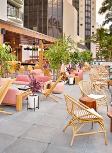 Open-air patio with colorful furniture, wooden chairs, and lush plants. Bar area with stools on the left, surrounded by tall buildings.