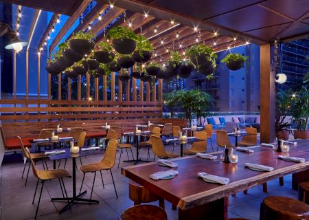 An outdoor restaurant patio with wooden tables, chairs, hanging plants, and string lights under a wooden pergola, creating a cozy atmosphere.