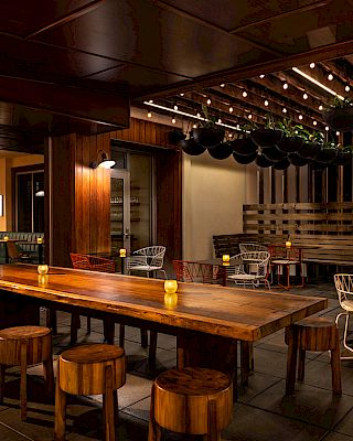 The image shows a cozy indoor restaurant or bar with wooden tables, stools, ambient lighting, and hanging plants for decoration.