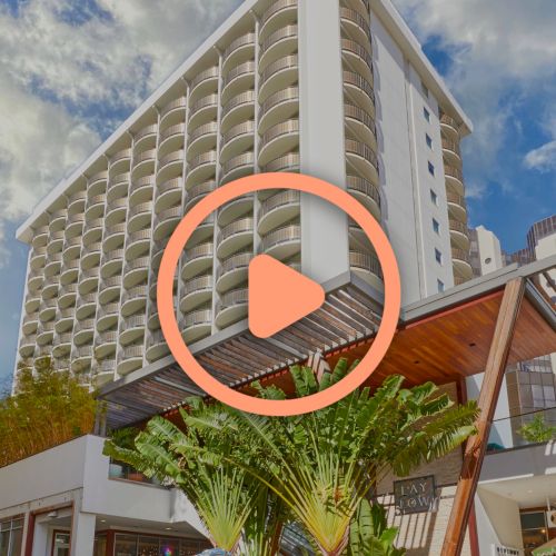 The image shows a tall building with a series of balconies, surrounded by trees, and a large play button overlay in the center.