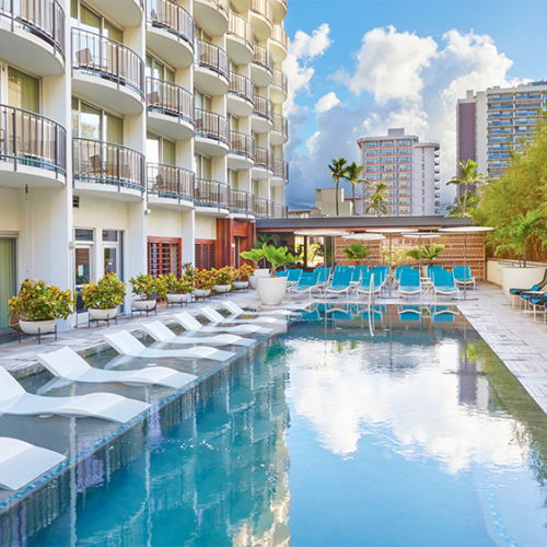 The image shows a hotel pool area with lounge chairs, surrounded by multi-story buildings and greenery under a partly cloudy sky.