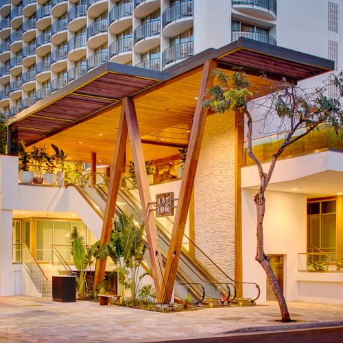 A modern building with a unique wooden entrance, glass railings, and balconies. It's surrounded by greenery and features outdoor lighting.