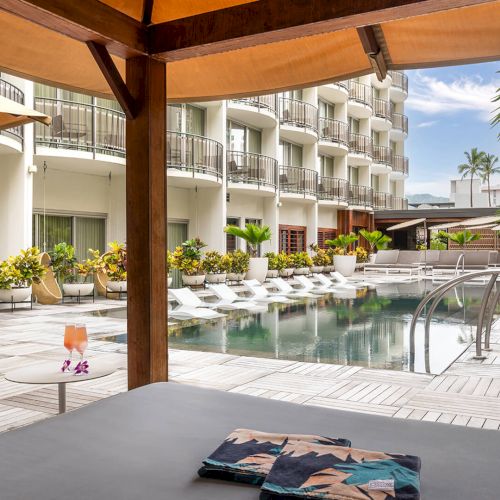 A poolside view with lounge chairs and plants, surrounded by a building and cabana, featuring a laid-back atmosphere.