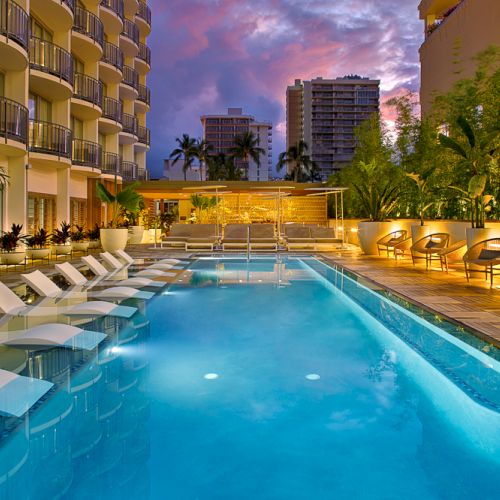 A modern outdoor pool area with lounge chairs, surrounded by buildings and greenery, under a vibrant evening sky with scattered clouds.