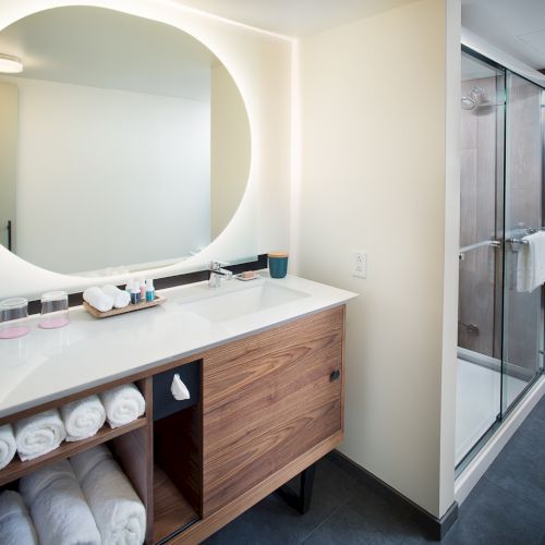 The image shows a modern bathroom with a round mirror, sink, wooden cabinet with towels, toiletries, and glass shower enclosure.