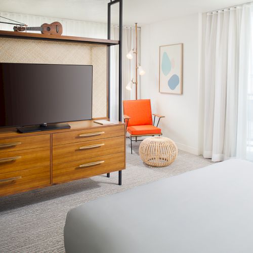 The image shows a modern hotel room with a TV on a wooden dresser, an orange chair, a round ottoman, and floor-to-ceiling curtains.
