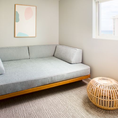 A cozy room with a gray sofa, a modern art piece on the wall, a round wicker ottoman, and a window providing natural light.