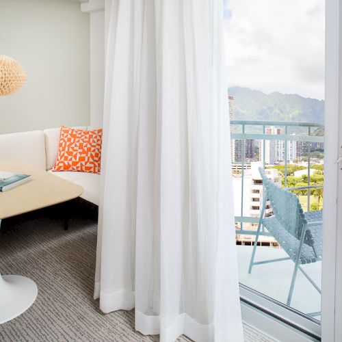 The image shows a cozy room with a small table, chairs, and a view of a balcony overlooking a cityscape and mountains.