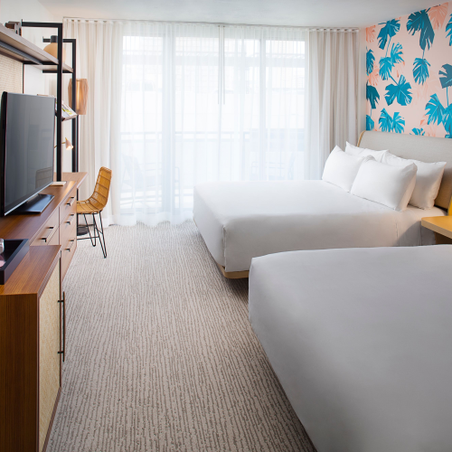 A modern hotel room with two beds, tropical wallpaper, a TV, a desk, and a view through sheer curtains.