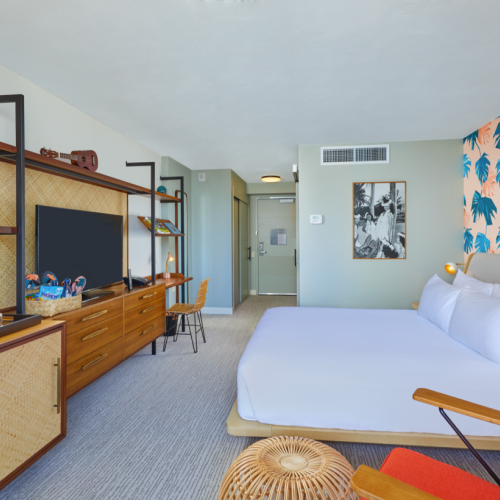 A stylish hotel room with a bed, tropical wallpaper, TV, and wooden furniture, featuring decorative items and a hallway door visible.