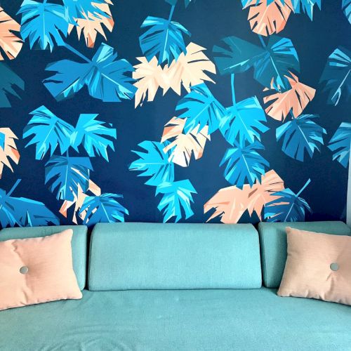 A light blue sofa with two pink cushions is in front of a vibrant wall featuring blue and peach leaf patterns.