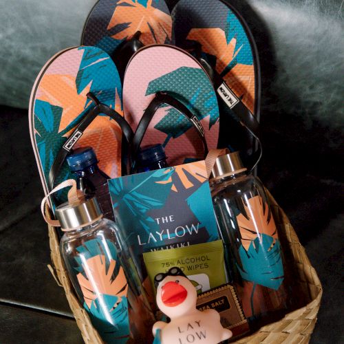A basket filled with flip-flops, water bottles, a booklet, and a rubber duck on a couch.