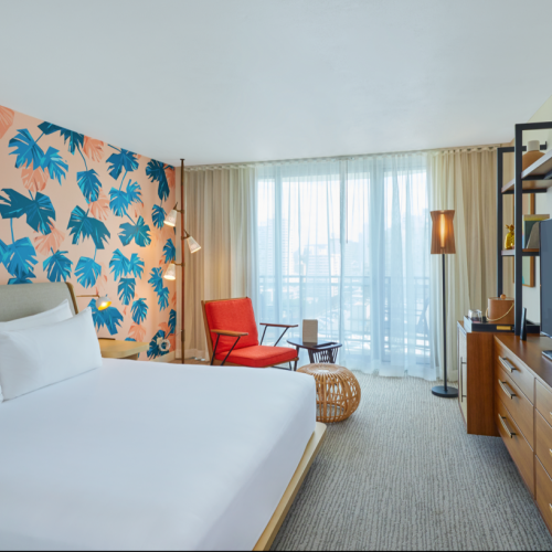 A hotel room with a tropical-themed wall, bed, TV, red chair, and large window leading to a balcony, creating a cozy atmosphere.