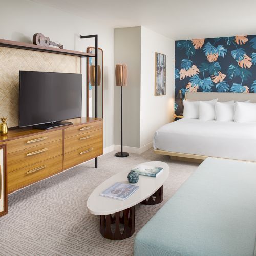 A modern hotel room with a king-size bed, TV, sofa, and tropical-themed decor on the wall behind the bed.