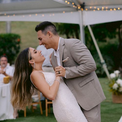 A couple is dancing under a tent, with string lights above them. The woman is in a white dress and the man in a suit, surrounded by greenery.