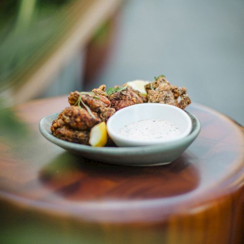 A plate of fried chicken pieces with lemon wedges and a bowl of creamy dipping sauce, set on a wooden surface outdoors.