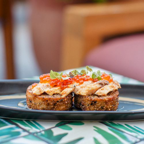 Two pieces of toast with toppings like sliced fish and herbs on a plate, set on a patterned table outdoors.