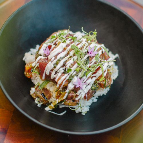 A bowl of white rice topped with various toppings, drizzled with sauce and garnished with sprouts and small edible flowers.